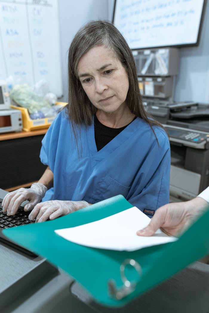 Nurse in scrub suit typing on keyboard and reviewing medical documents in a clinic setting.